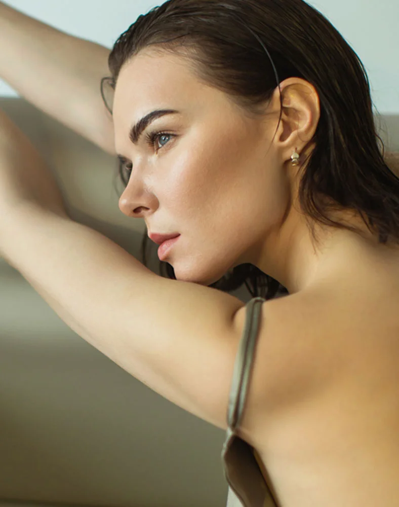 Close-up profile view of a light-skinned woman with wavy, dark brown hair that appears wet or styled. She has a smooth complexion with subtle blush on her cheeks and light blue eyes gazing off to the side. Her expression is neutral. One bare arm is loosely draped in view, showing light-toned skin. A thin, pale olive-colored strap from a sleeveless top or dress crosses her shoulder. The background is a muted sage green or beige, and the image is softly lit with natural light. - BBL Laser Treatment in Marina Del Rey, CA