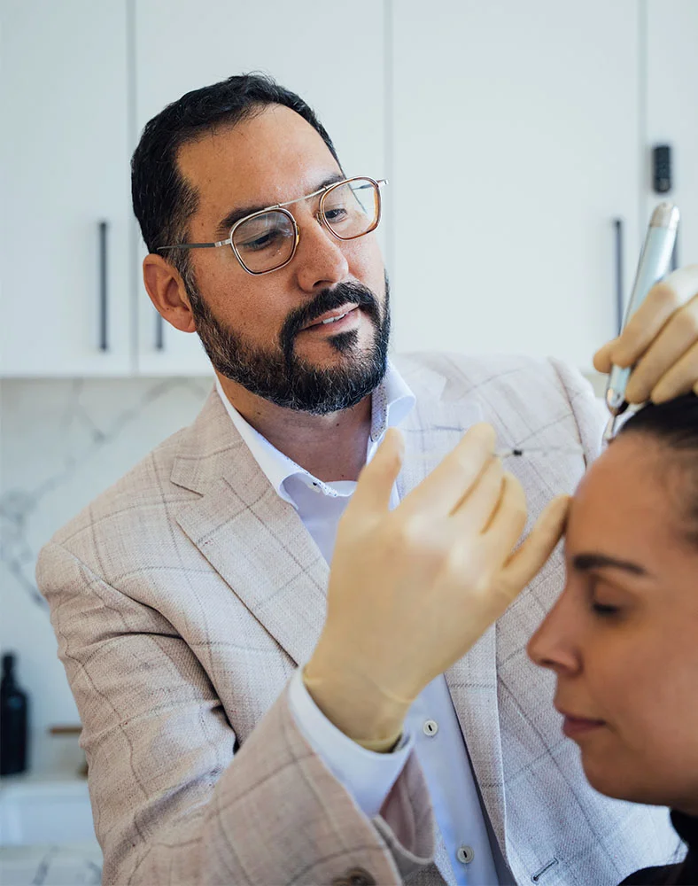 Dr. Macias, a light-skinned man with a beard and glasses, wears a light beige patterned suit jacket, a white shirt, and pale yellow gloves. He is focused intently on a woman in front of him, whose eyes are closed. His hands are positioned near her forehead, suggesting a medical or cosmetic procedure. The background features light-colored cabinets or walls, and the lighting is bright and clinical. - Botox and Wrinkle Relaxers in Marina Del Rey, CA