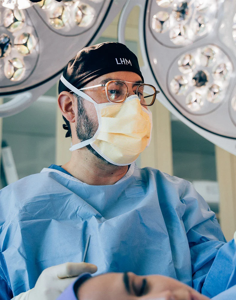 A close up shot of Dr. Macias preforming surgery on a client under large surgical lights. He wears scrubs, mask and gloves as his patient, slightly seen in shot, lays below with her eyes closed. - Buccal Fat Removal in Marina Del Rey, CA