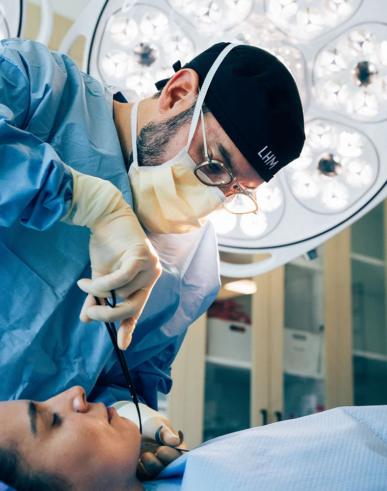 A close-up of Dr. Macias performing surgery, wearing a blue gown, black cap, and yellow mask. Focused hands handle instruments over a patient draped in light blue surgical sheets. The sterile operating room and overhead surgical lights are visible, emphasizing precision and care. - Chin Augmentation in Marina Del Rey, CA