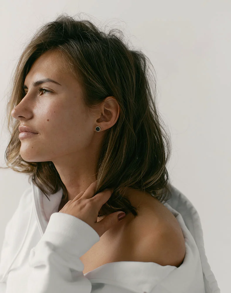 A close-up, slightly low-angle shot of a light-skinned woman with shoulder-length brown hair. She is seen from the mid-chest up, gazing softly off to the viewer’s right with a neutral expression. A small dark stud earring adorns her ear. She wears a loose off-white top, with one hand gently touching her neck near the shoulder. Soft, diffused lighting casts a gentle glow on her face and shoulders against a plain, light neutral background. - Cryo Correct in Marina Del Rey, CA