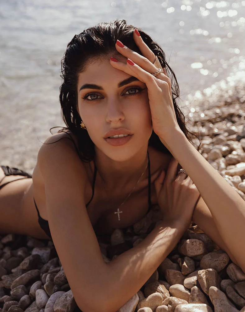 A close up shot of a woman with dark brown hair laying on a rocky beach wearing a black bikini. She holds one of her hands holding her other arm while the other grazes her forehead and hair. - Dermal Fillers in Marina Del Rey, CA