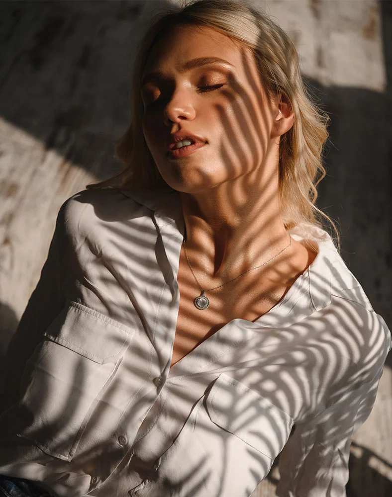 A close up shot of a woman laying of a wood floor glancing up at the camera with her eyes closed as the palm tree shadows hit her face. She wears a white button up and long blond curly hair. - Dermaplane in Marina Del Rey, CA
