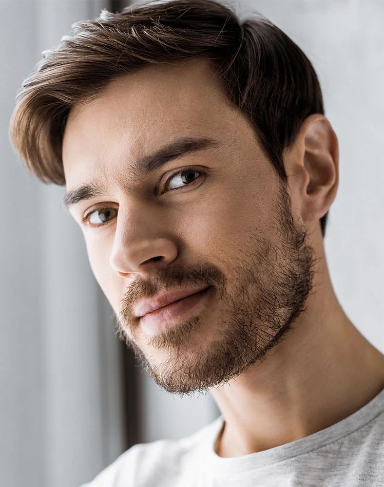 A close up shot of a man wearing a white t-shirt glancing into the camera. He has short brown hair and a well groomed beard and mustache. - Ear Surgery for Men in Marina Del Rey, CA