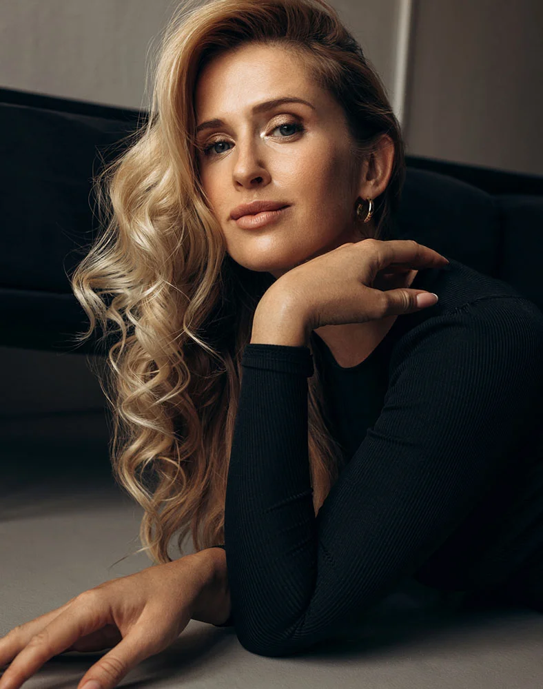 A light-skinned woman with shoulder-length wavy blonde hair lies on a light gray surface, wearing a black fitted long-sleeve top. She looks calmly and directly at the camera with a serene expression. Her right arm rests gently on her face, and gold hoop earrings are visible. The background is a muted gray with a dark, blurred chair or sofa. Soft lighting highlights her features in a relaxed, sophisticated portrait. - Eyelid Surgery in Marina Del Rey, CA
