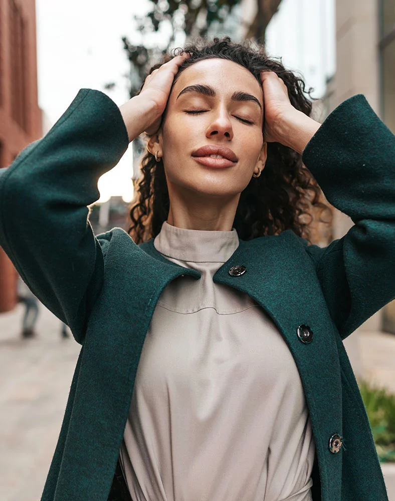 A close-up captures a woman with curly, dark brown hair. She wears a dark teal, wool-like coat over a light beige or taupe mock-neck top. Her eyes are closed and her hands softly rest on her head, conveying a relaxed and serene expression. The background is an out-of-focus urban scene with buildings and greenery, suggesting an outdoor daytime setting. - Forever Clear in Marina Del Rey, CA