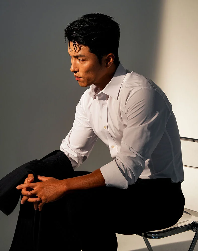 A man with neat dark hair sits in a chair, shown in a three-quarter view. He wears a white shirt and dark pants, his arms resting on his lap as he gazes downward contemplatively. Soft studio lighting highlights him against a muted gray background, creating a professional atmosphere. - Fue Hair Restoration in Marina Del Rey, CA