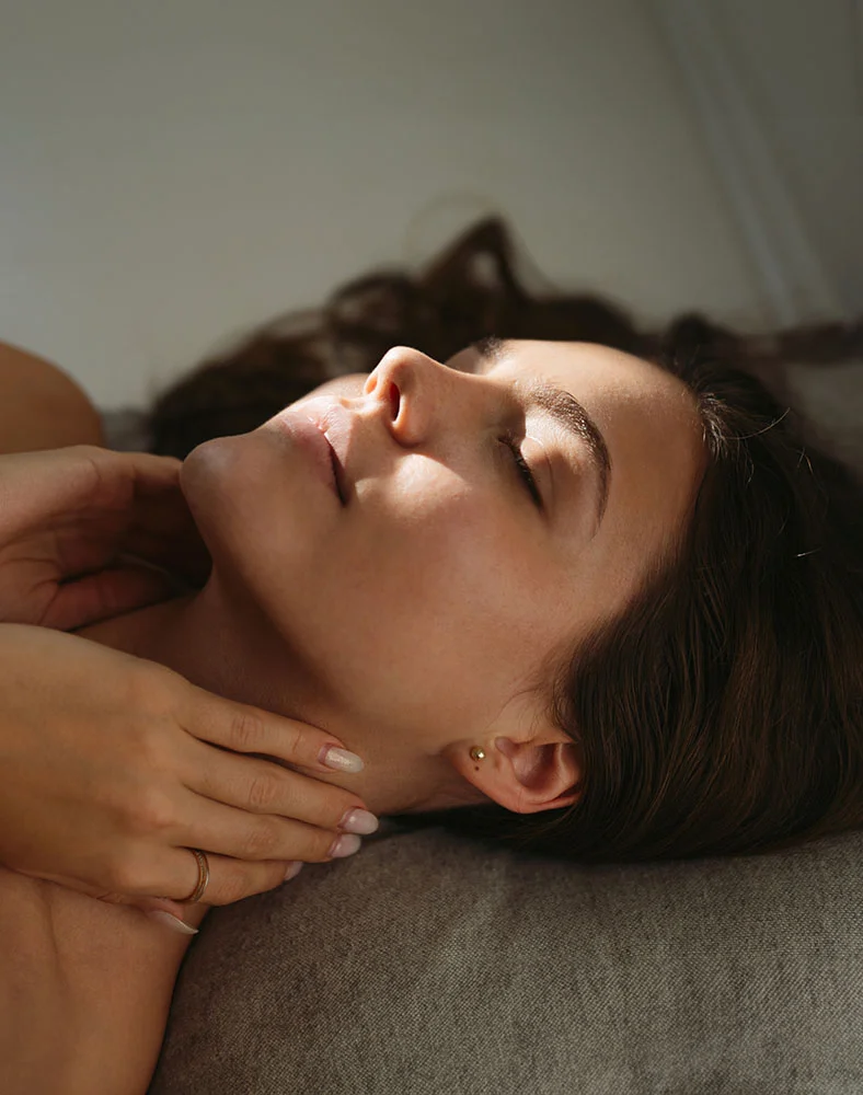 A light tan woman with long, dark brown hair lies on her back, eyes closed, head tilted, with a relaxed and peaceful expression. Small gold stud earrings are visible. Her hands rest gently on her neck and upper chest on a grey fabric surface, revealing a delicate ring on her ring finger. Soft, natural light casts gentle shadows, focusing on her face and neck, creating a serene and calming aesthetic. - Halo in Marina Del Rey, CA