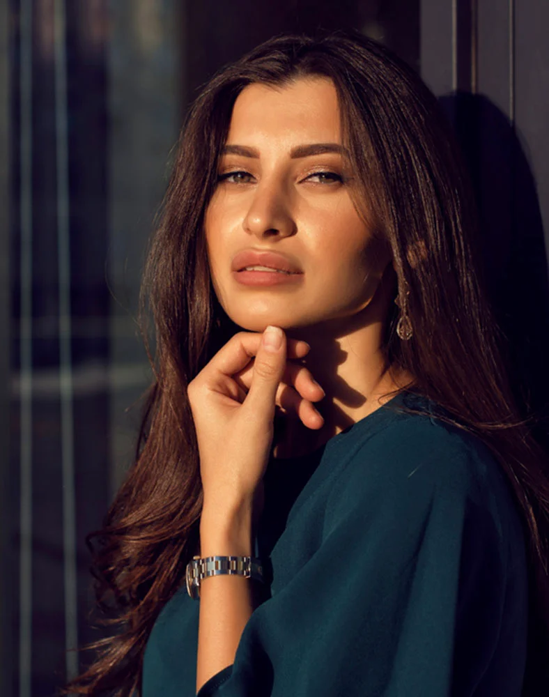 A close-up portrait shows a light-skinned woman with long, dark brown hair and soft, natural makeup. She has a thoughtful expression, looking directly at the camera with light hazel or brown eyes and rosy pink lips. She wears a teal or deep emerald green top, with a silver or stainless steel watch on her right wrist and a small gold earring. The dark, out-of-focus background with blurred patterns suggests a window or doorway with soft sunlight. - Hydrafacial in Marina Del Rey, CA