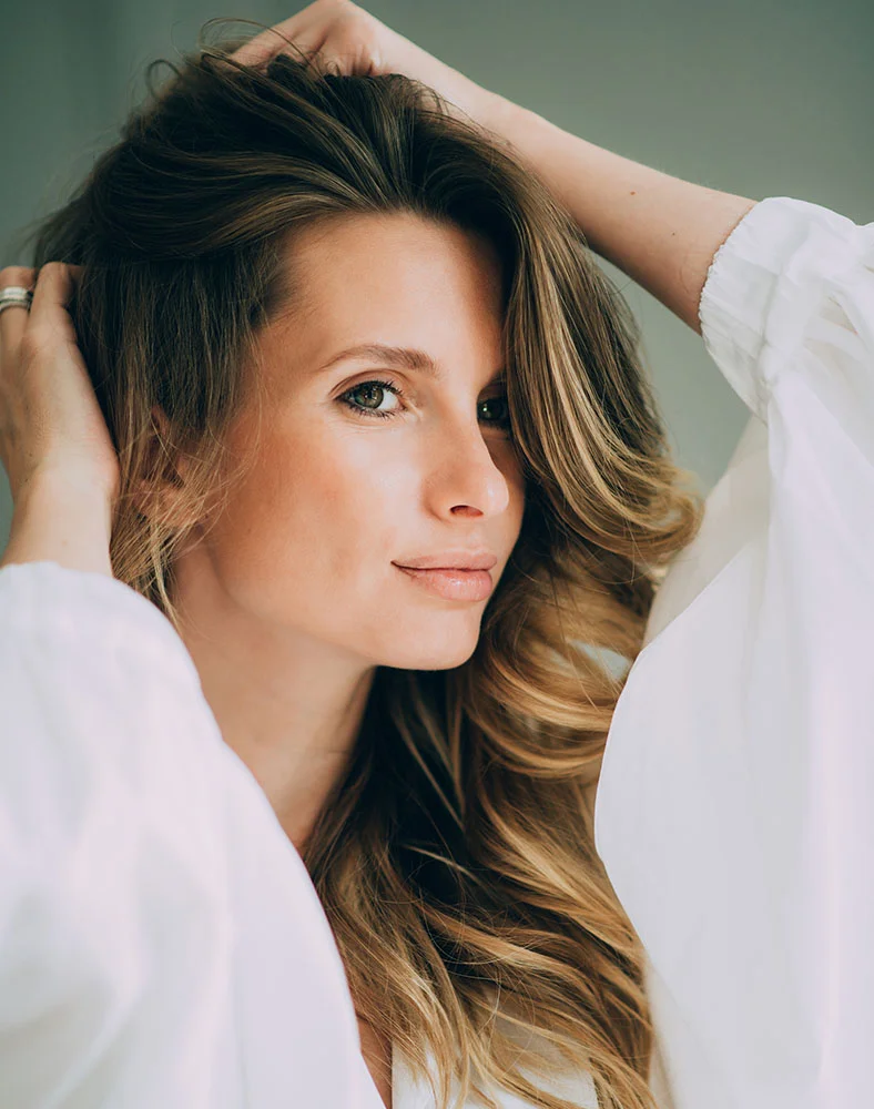 A light-skinned woman with shoulder-length, loosely waved, light brown hair, parted on the side, is shown from the mid-chest up. She wears a loose-fitting, off-white, long-sleeved top. Her expression is serene and slightly thoughtful, with her gaze directed to the viewer's right. She holds her hair with both hands close to her head. The background is a muted, light sage-green. The image is well-lit and focuses on her facial features and hair. - Keravive in Marina Del Rey, CA