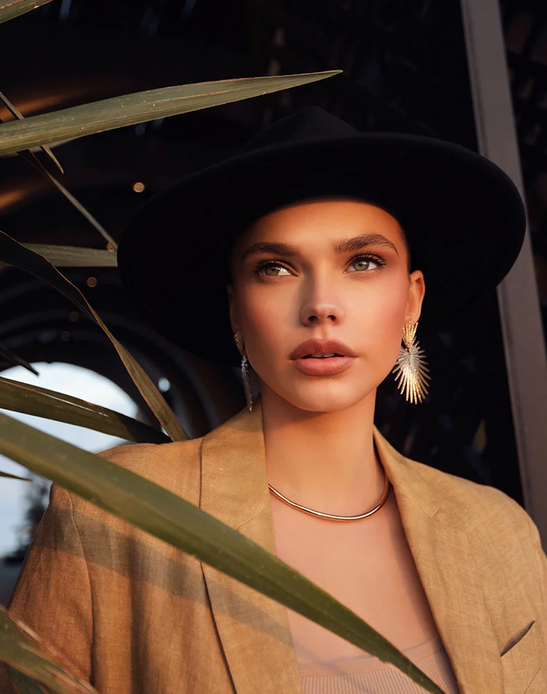 A young woman with light skin and light eyes gazes slightly upward and to her right. She wears a black fedora, a tan blazer over a light-colored top, a gold choker, and large, ornate gold and silver earrings. Lush green foliage frames the foreground, and the background is dark and out of focus. - Lip Lift in Marina Del Rey, CA