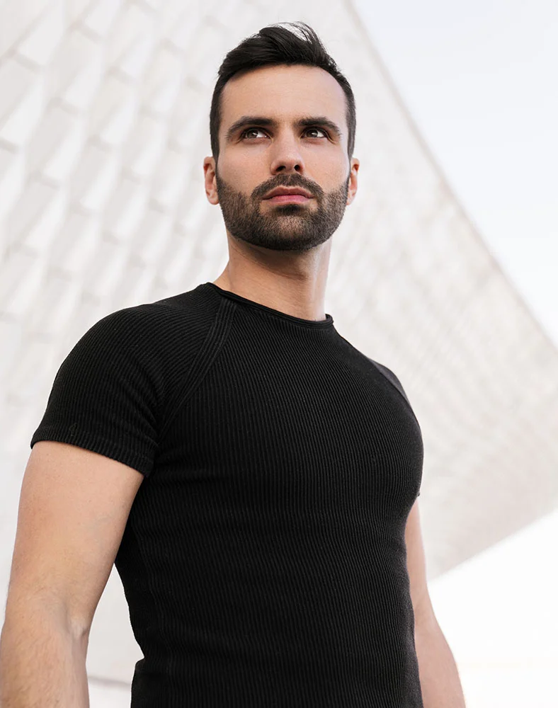 A medium close-up shows a man with short, dark brown hair and a trimmed beard, looking upward and slightly to his right with a neutral or thoughtful expression. He wears a close-fitting, black ribbed or knit short-sleeved t-shirt, revealing his arms, shoulders, and upper body. The out-of-focus background is a light gray or white architectural structure with abstract shapes and angles. The lighting emphasizes his features and the clothing's texture. - Men Body Contouring in Marina Del Rey, CA