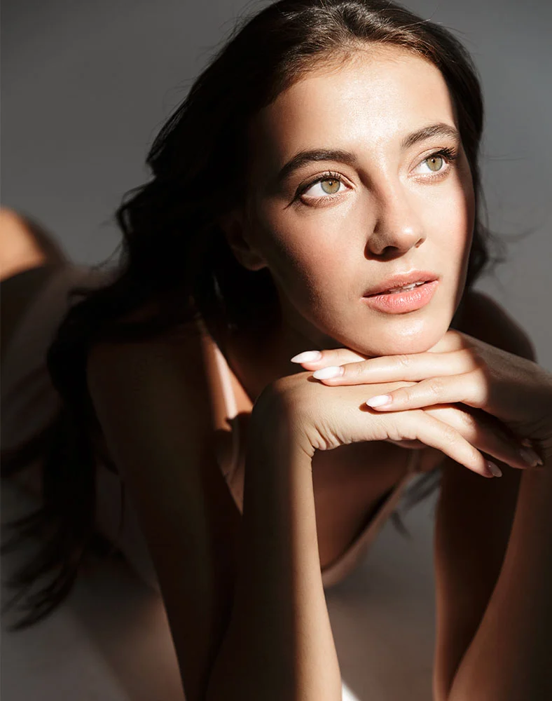 A woman with long, dark brown hair lies on a light gray surface, her head slightly elevated, looking to the upper right with a serene, thoughtful expression. Her light, manicured hands rest lightly on her face, supporting her chin and side of her head. Dramatic lighting from above and slightly left creates strong shadows and highlights on her features and soft shadows on her arm and hair. She wears a light beige or pinkish-tan top subtly contrasting with her skin tone. - Microdermabrasion in Marina Del Rey, CA