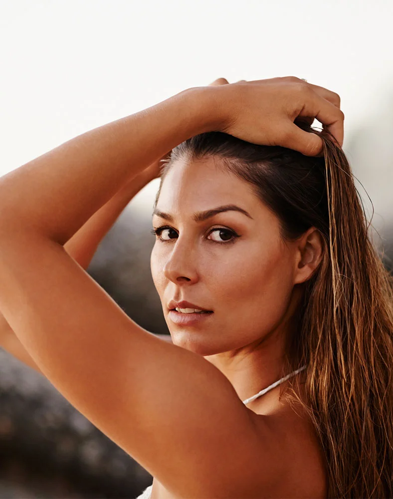 A light-skinned woman with long, dark brown hair pulls her hair back with her hands, looking directly at the camera with a neutral expression. She wears a simple white necklace. The background is a blurred mix of light and dark, suggesting an outdoor setting with rocks. - Mini Facelift in Marina Del Rey, CA