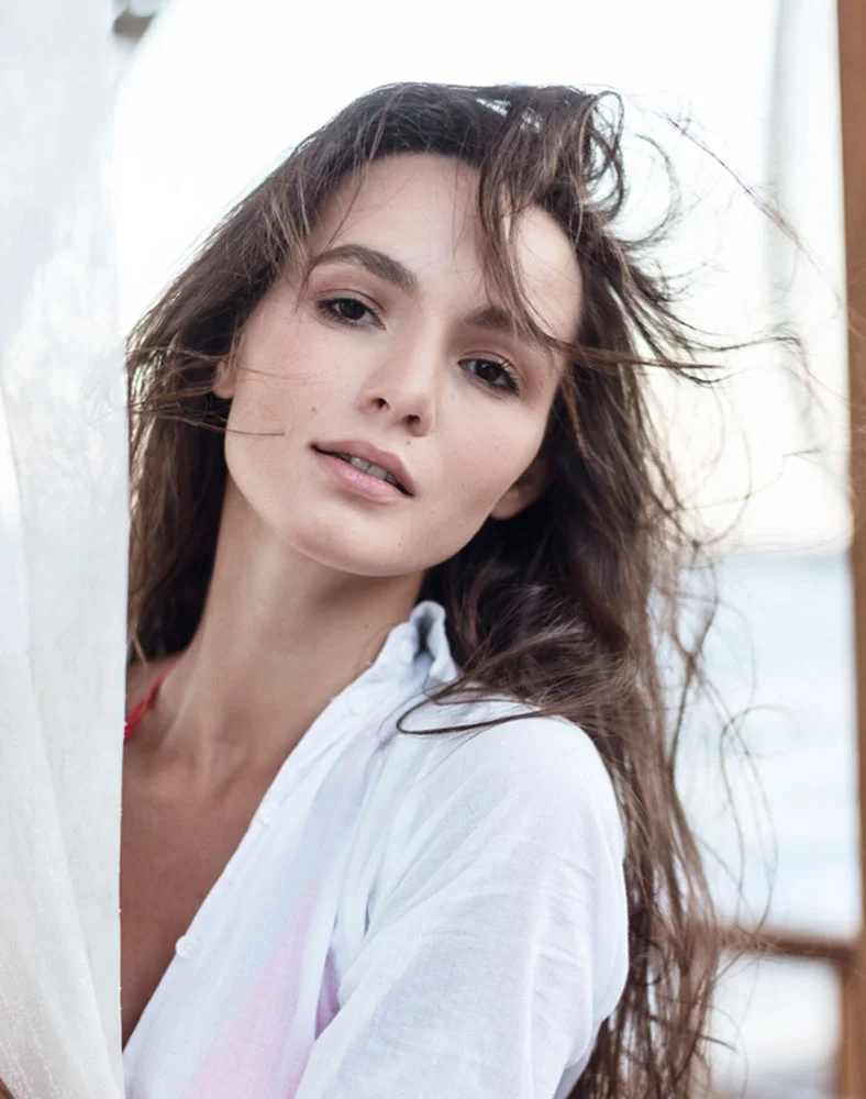 A light-skinned woman with long, dark brown hair flowing around her face looks directly at the viewer with a neutral expression. She wears a loose-fitting white cover-up or shirt over a red top, hinting at a beach or poolside setting. White sheer fabric is visible to her left, and a blurred light background with a hint of water is behind her. - Renuvion for Skin Tightening in Marina Del Rey, CA