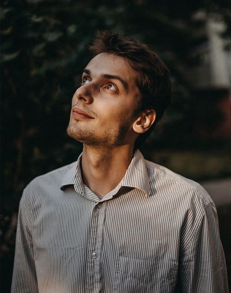A close-up portrait of a light-skinned man with short dark hair and a light beard, looking upwards and slightly to his left with a thoughtful expression. He wears a light-colored, striped button-down shirt. The background is dark and out of focus, with hints of green foliage. - Rhinoplasty for Men in Marina Del Rey, CA
