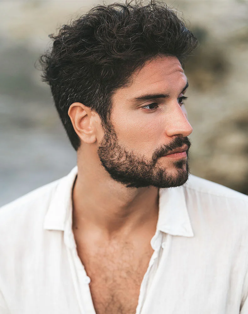 A close-up side profile of a light-skinned man with dark, curly hair and a full beard, looking to his right with a neutral expression. He wears a light-colored, unbuttoned collared shirt, revealing some chest hair. The background is blurred and light, suggesting an outdoor setting with rocks. - Septoplasty in Marina Del Rey, CA