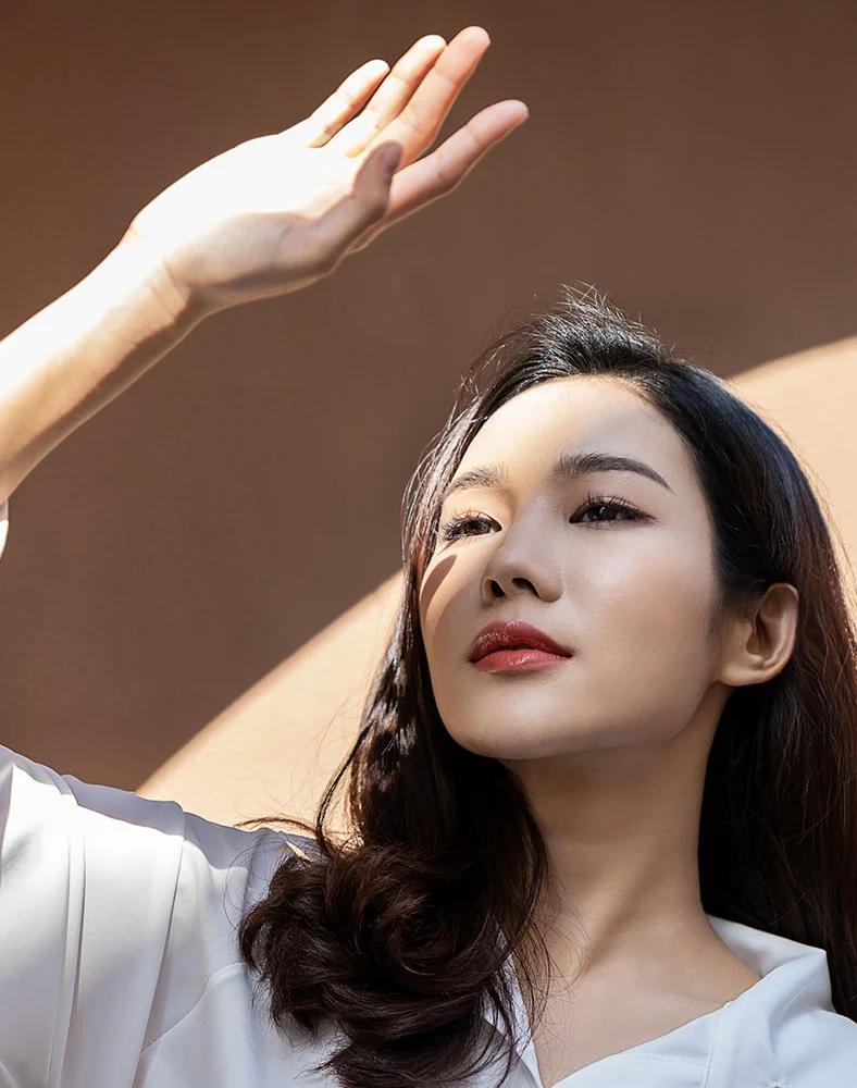 A close-up portrait of a light-skinned woman with long, dark brown hair looking upwards and to her right, shielding her eyes with her right hand. Sunlight casts strong, angular shadows across her face and a plain, warm brown background. She wears a white top and has light makeup with red lips. - Thread Lift in Marina Del Rey, CA